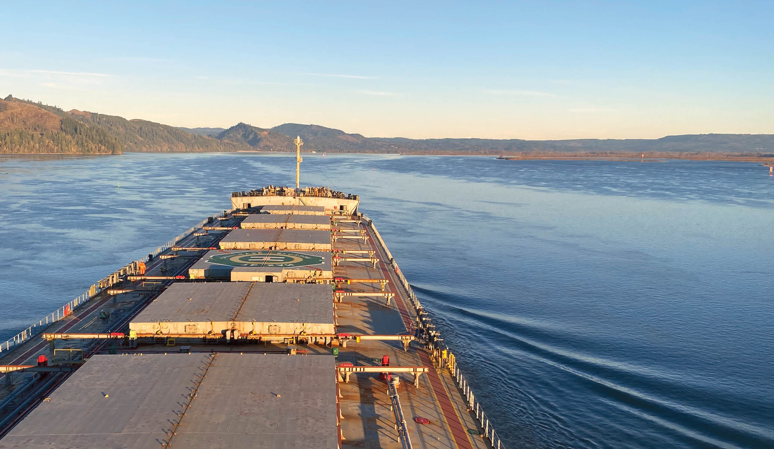 A photo of a bulk carrier ship, taken from the bridge, with the landscape of the Columbia River beyond the prow of the vessel