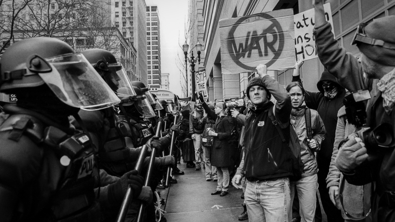 A row of police in riot gear faces a line of protesters in downtown Portland. One holds a sign reading "no war" and looks at the camera.