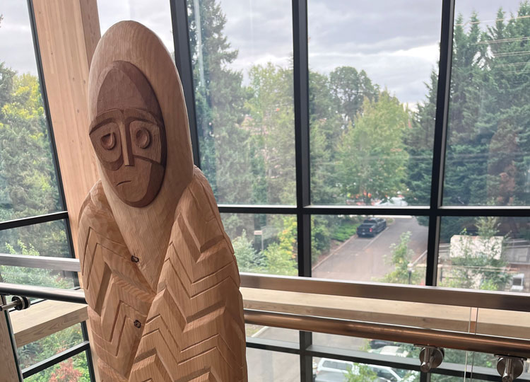A wooden carving of a person situated on a high floor with trees visible through windows outside