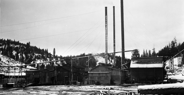 A black-and-white photo of the Kinzua Pine Mill in operation. The building has three smokestacks and is surrounded by snow.