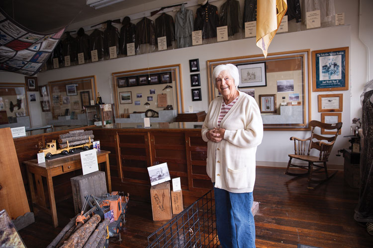 A photo of Pat Hyatt, a white-haired woman, standing in a room full of historical artifacts