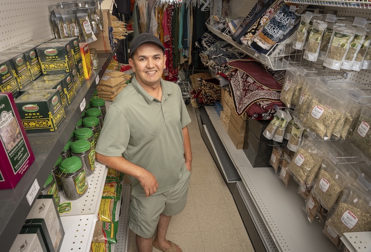 Mohammad Yonus Ghaznawi stands in an aisle of his grocery store, Global Mart. He is surrounded by teas, spices, and rugs.