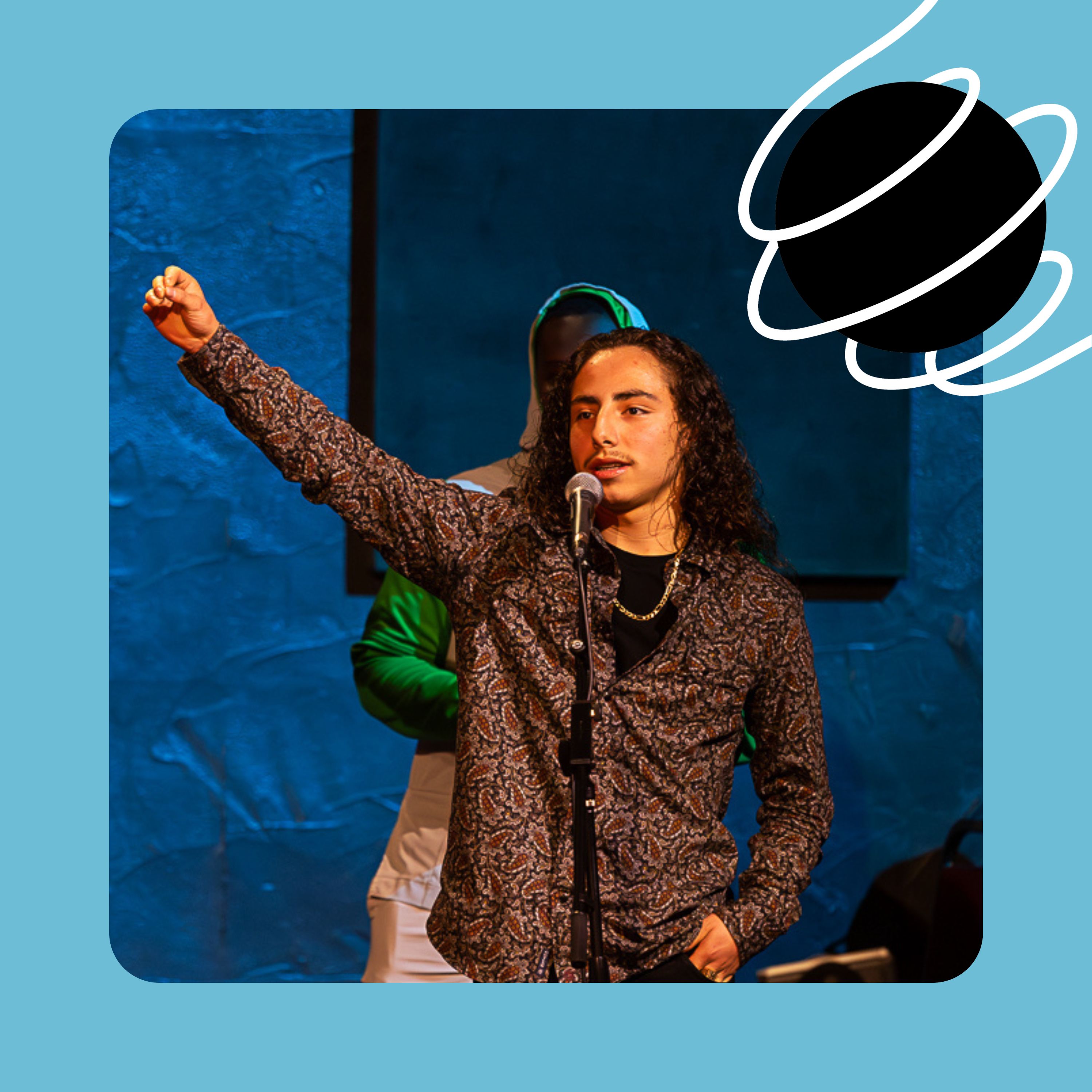 Malaki, a young man with long hair and a moustache wearing a paisley-print shirt, stands at a microphone with his right fist raised in the air.